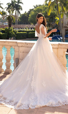 A woman in the Long Wedding Dress 11134 by Primavera—an ivory A-line gown with lace details and cape short sleeves—stands by a stone railing, overlooking a pool with palm trees and a "PRIMAVERA" sign in the background.