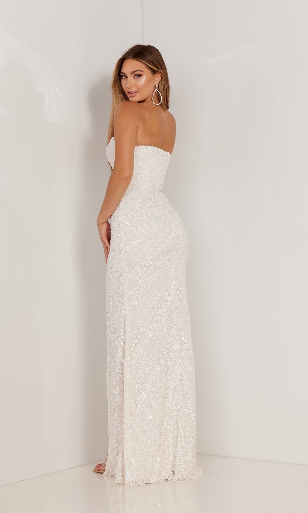 A woman stands facing away, wearing the Aleta Long Prom Dress 1118—a strapless, floor-length white gown with geometric sequin patterns.
