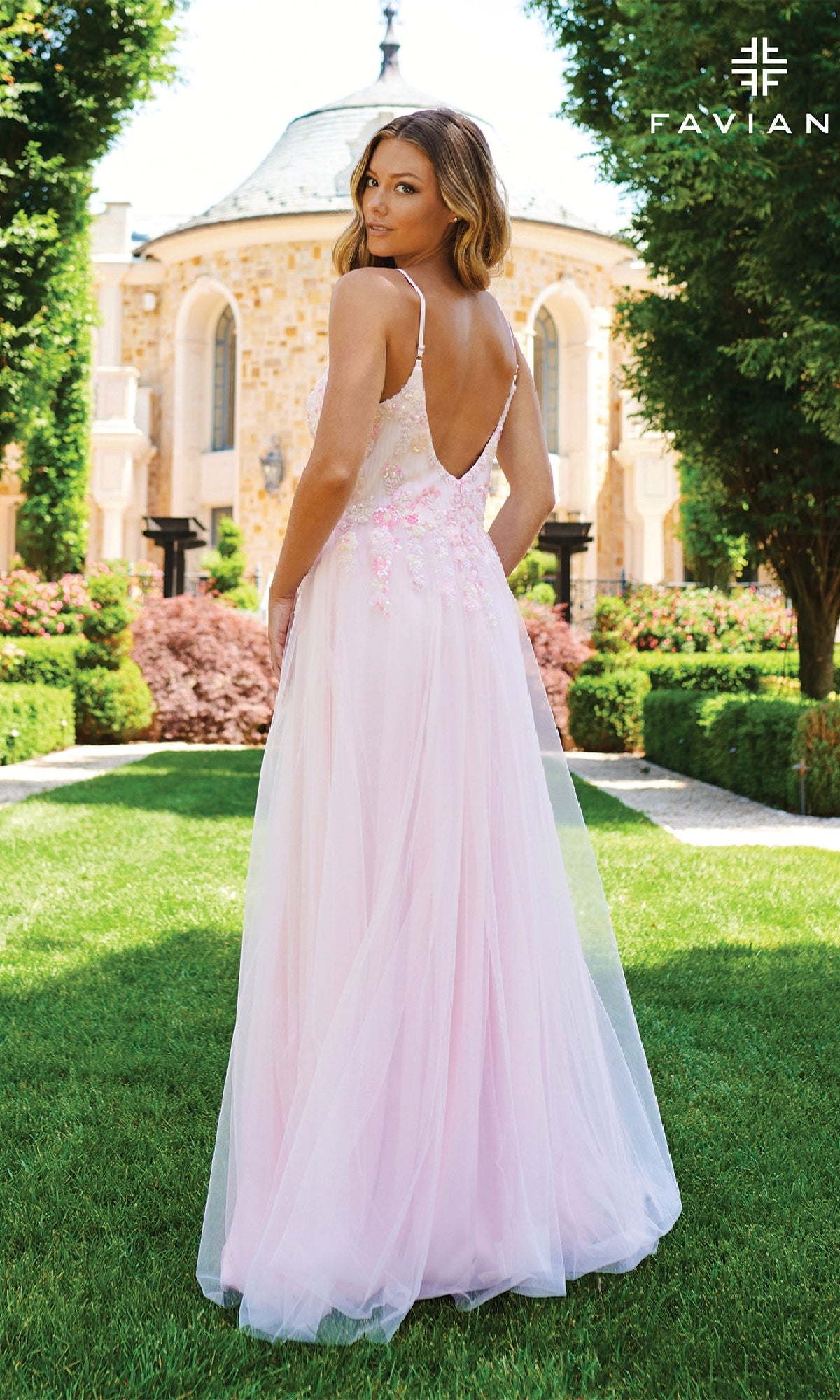 A woman stands outdoors in front of a stone building, wearing the Faviana 11232—a long pastel A-line prom dress with a v-neckline, thin straps, backless design, and delicate floral details.