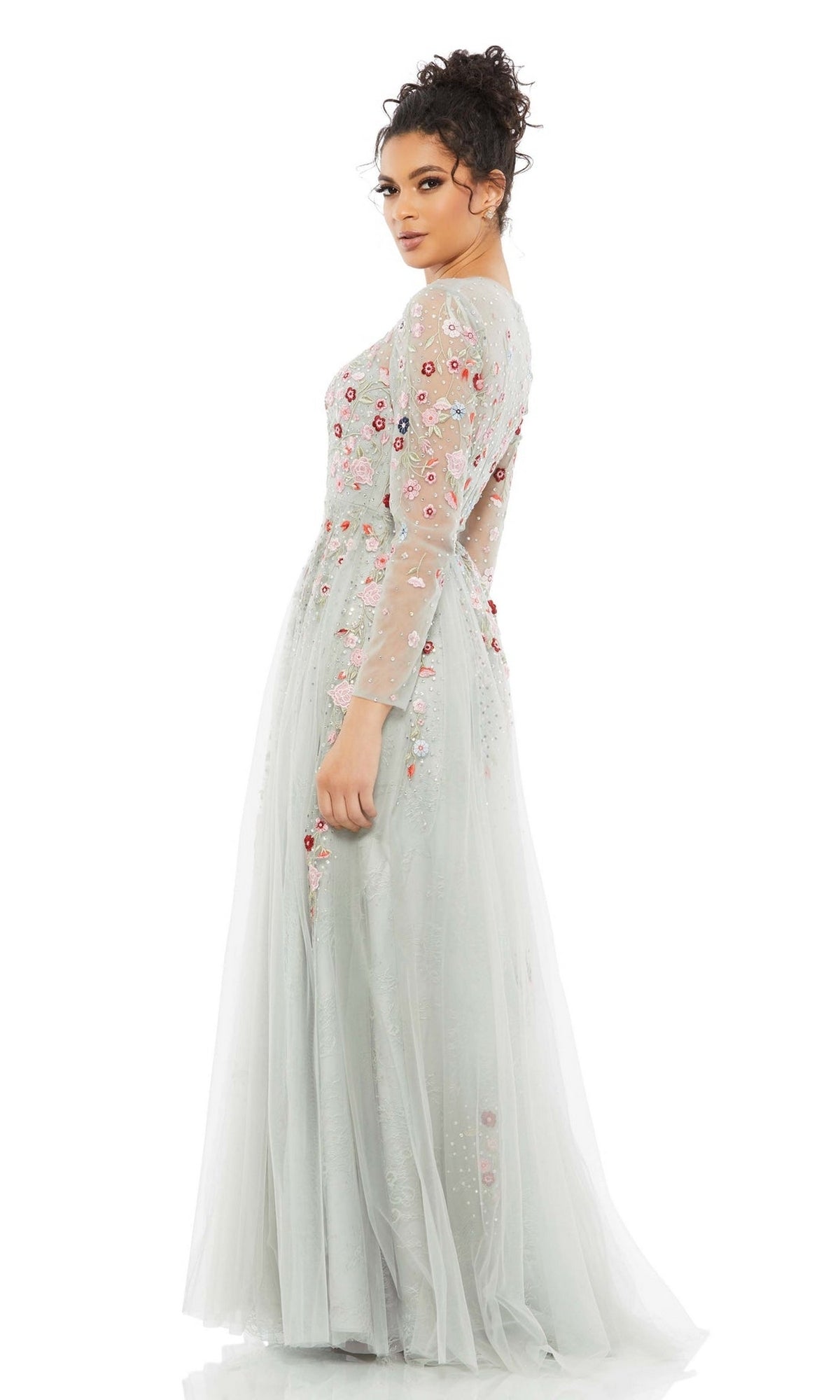 A woman wears the Long Formal Dress 11241 by Mac Duggal—a light blue A-line gown with sheer long sleeves and colorful floral embroidery—standing away and glancing over her shoulder.