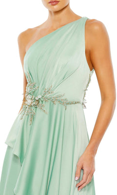 A woman wearing the Long Formal Dress 11262 by Mac Duggal—a light green, one-shoulder gown with an embellished waist, jeweled floral details, and a draped, flowing skirt.