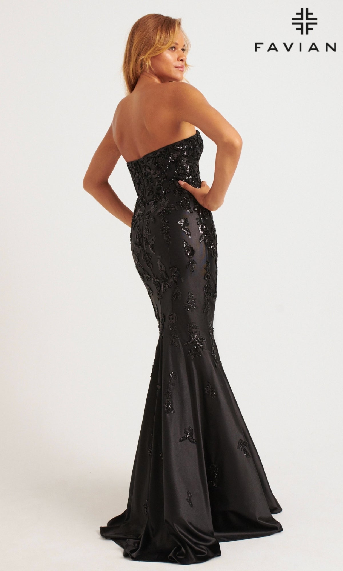 A woman poses with her back turned in the Faviana 11271, a strapless fitted black mermaid prom dress adorned with sequin embellishments. The gown elegantly flares at the bottom and 