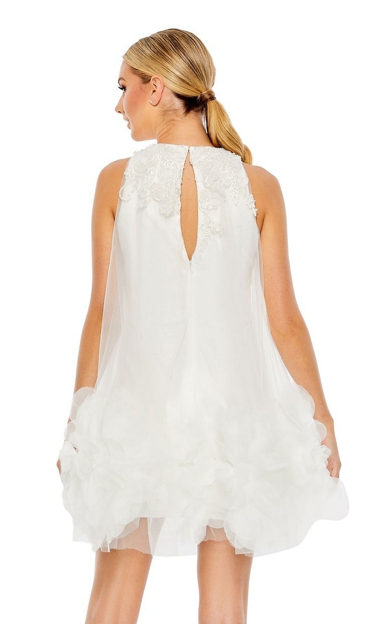 A woman stands facing away, her hair in a low ponytail, wearing the Mac Duggal Short Homecoming Dress 11294—an A-line, sleeveless white party dress with floral appliques and a keyhole back.