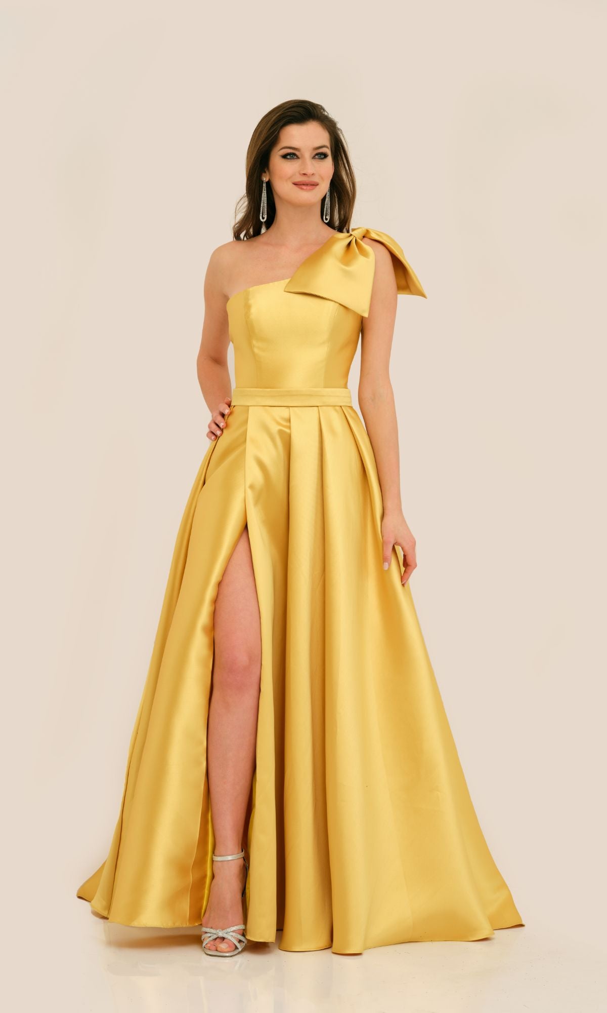 A woman wears the Long Formal Dress 11337 by Dave and Johnny—a floor-length, one-shoulder gown with a bow detail and thigh-high slit—paired with silver heels and earrings, standing against a plain background.