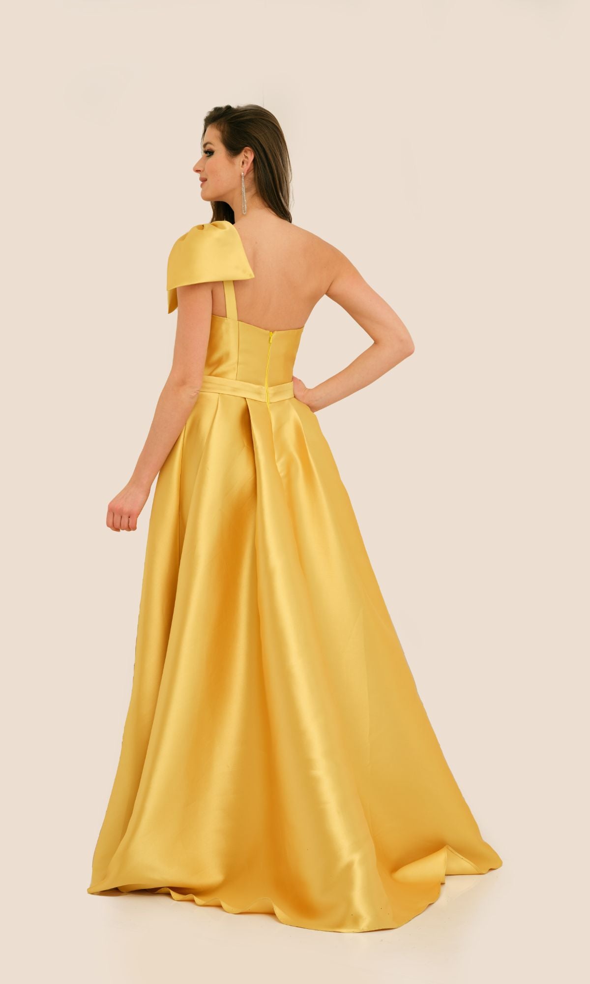 A woman faces away against a beige background, wearing the Long Formal Dress 11337 by Dave and Johnny—a floor-length, yellow satin A-line dress with pleats.