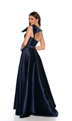 A woman poses against a white background in the Long Formal Dress 11337 by Dave and Johnny, a floor-length dark blue satin one-shoulder ball gown with a fitted bodice and large shoulder bow, looking over her shoulder.