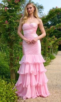 A woman stands outdoors in a light pink Faviana 11419 long prom dress featuring a strapless sweetheart neckline, ruched bodice, and layered ruffle skirt, surrounded by lush greenery and blooming flowers.