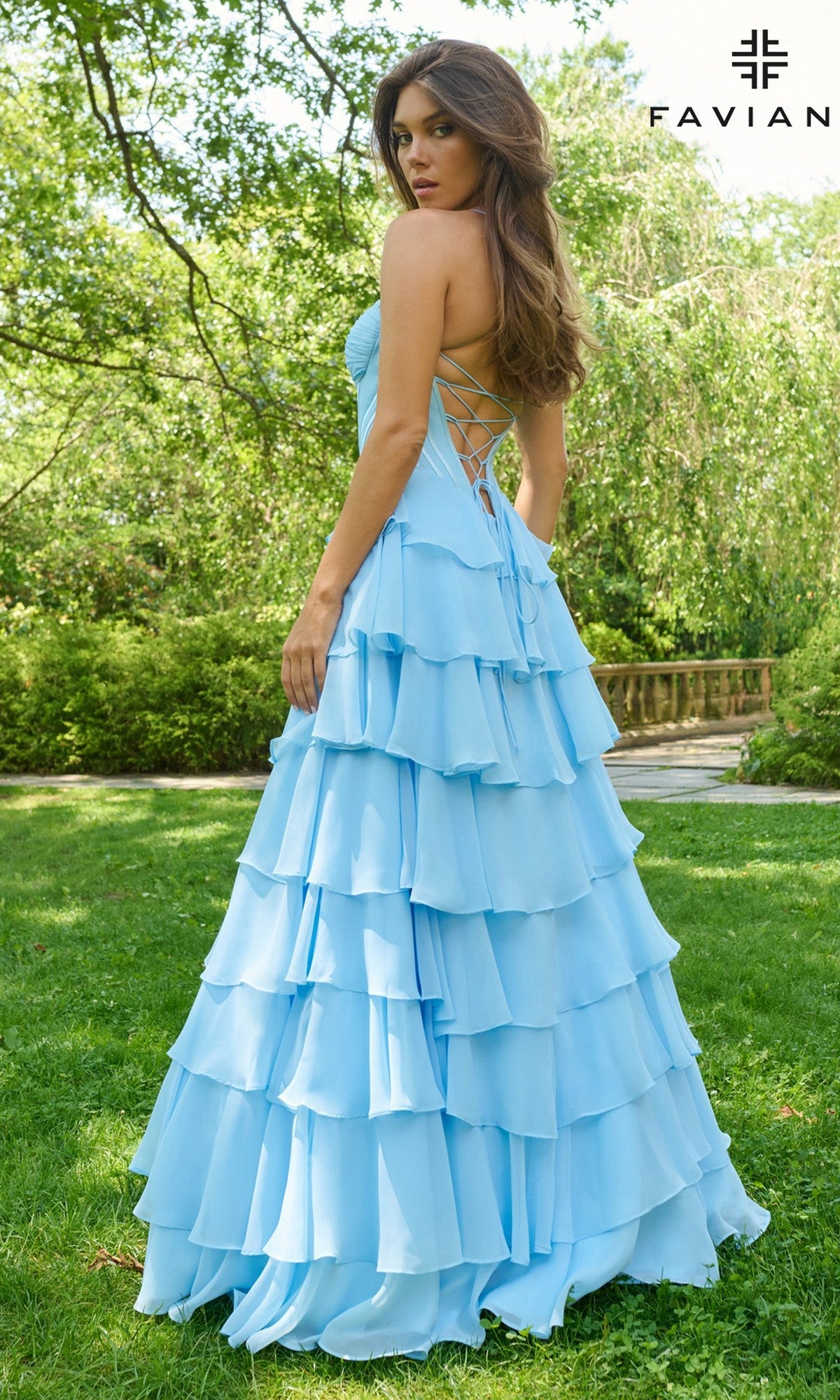 A woman stands outdoors on grass in a light blue Long Prom Dress: Faviana 11420, featuring chiffon fabric, a tiered skirt, backless design, and lace-up corset bodice. Trees are in the background; the Faviana logo appears in the top right corner.
