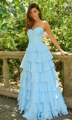 A woman stands outdoors by a stone railing in the Faviana 11420 long prom dress, a light blue chiffon gown with a corset bodice, tiered ruffle skirt, thin straps, and fitted silhouette, surrounded by greenery.