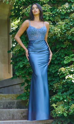 A woman wearing the Faviana 11433 Sheer Lace Corset Long Prom Dress stands on stone steps before green foliage. The dress is blue, fitted, sleeveless, with a beaded corset-style bodice.