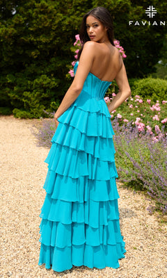 A woman stands outdoors on a gravel path in a Faviana 11436 Strapless Long Ruffled Prom Gown—a tiered turquoise dress—surrounded by greenery and pink flowers.