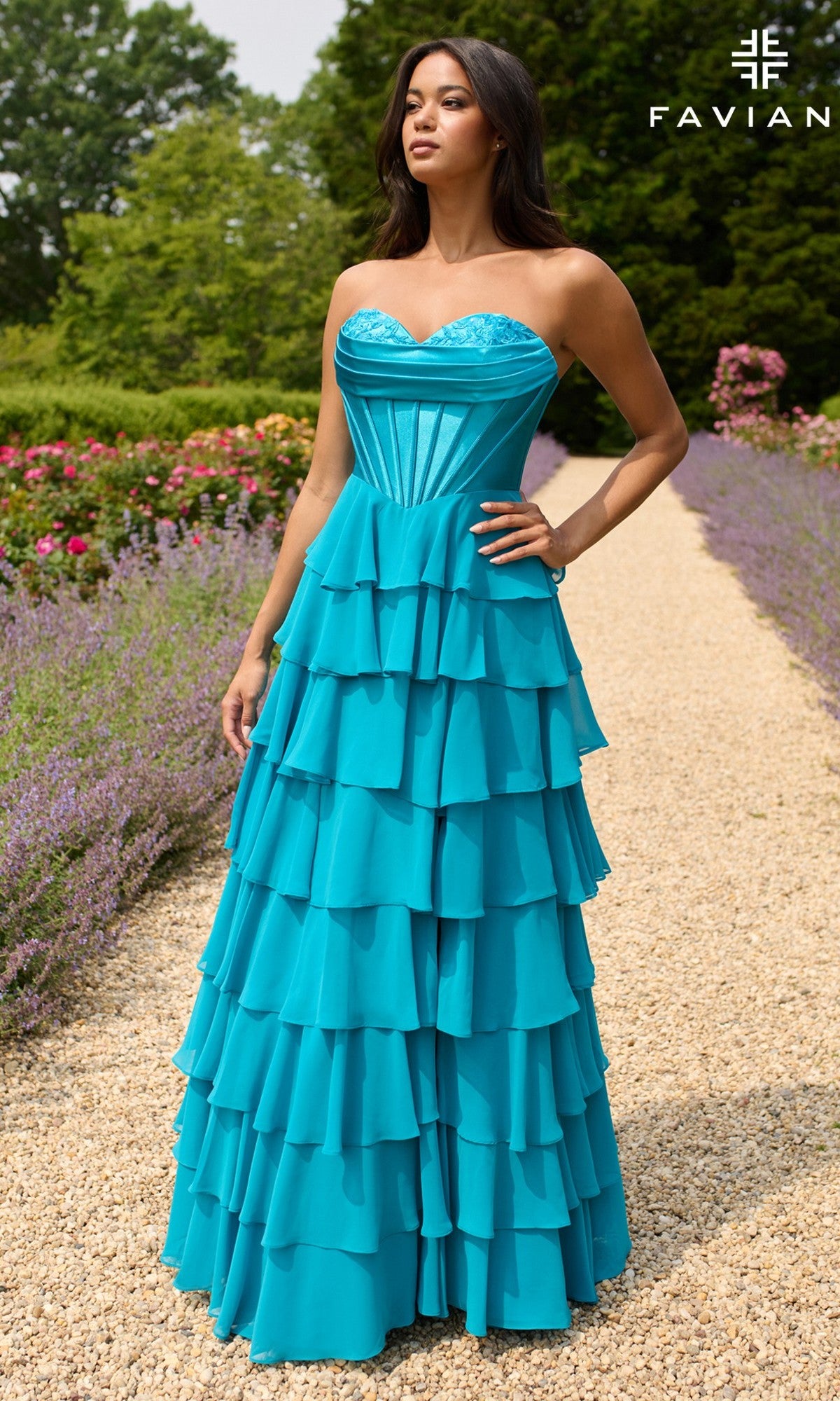 A woman stands on a garden gravel path in the Strapless Long Ruffled Prom Gown: Faviana 11436, a turquoise dress, surrounded by vibrant flowers and lush greenery.