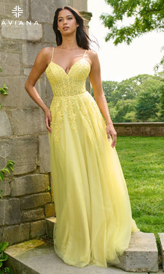A woman stands outdoors by a stone wall, wearing the Faviana 11438 long prom dress with thin straps, floral details, and an elegant yellow a-line tulle skirt.