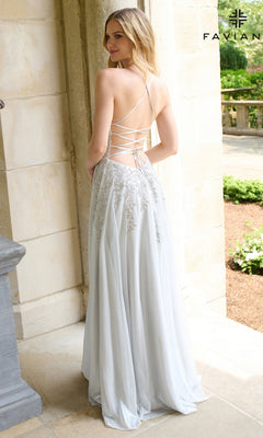 A woman stands outdoors, facing away from the camera, wearing the Long Prom Dress: Faviana 11449 in light blue with lace-up straps and embroidered details. The scene features stone columns and lush greenery.