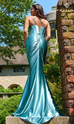 A woman in the Faviana 11454 long prom dress, featuring shiny teal satin and a lace-up back, stands outdoors by a brick pillar and greenery with a large building behind her.