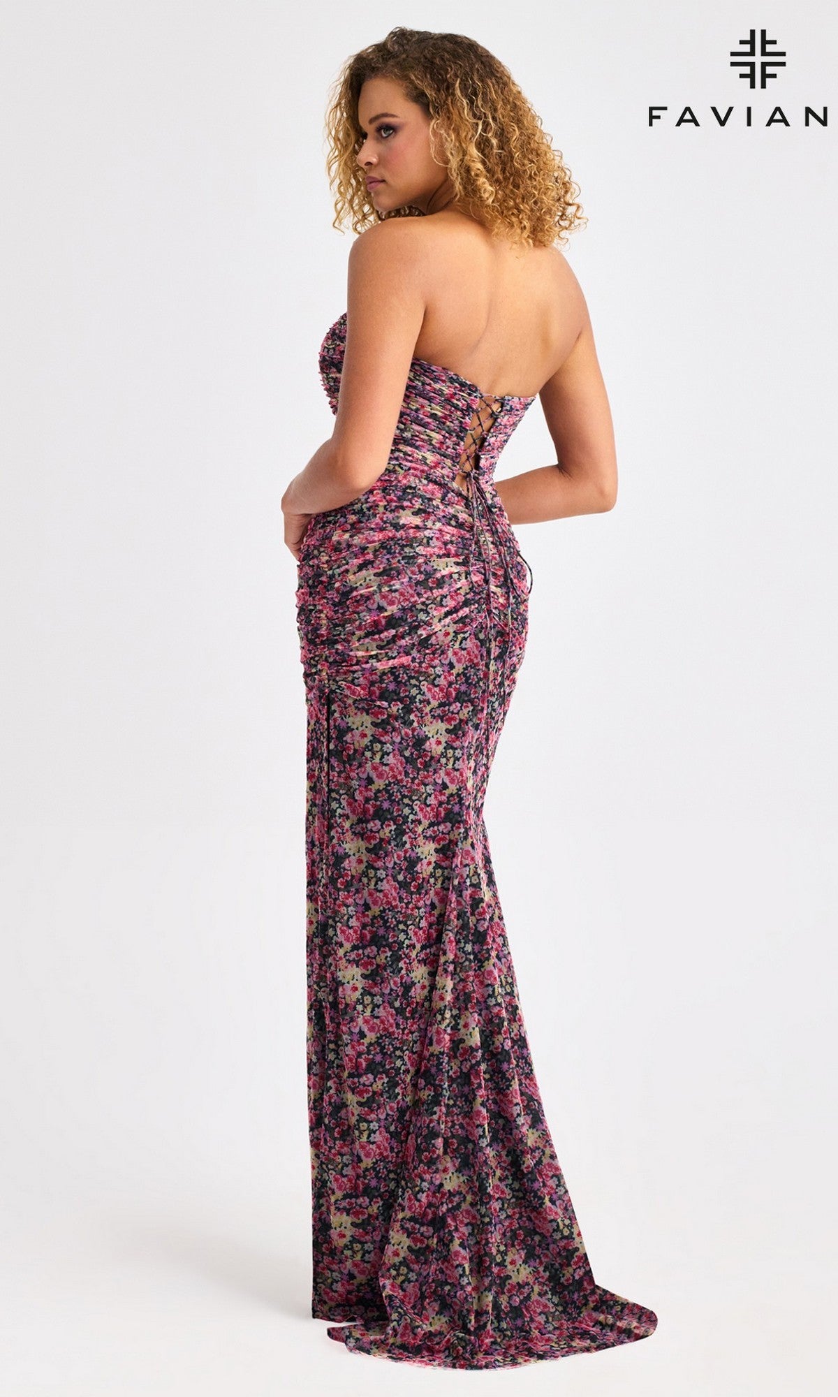 A woman in the Faviana 11461 long prom dress featuring a strapless neckline, floor-length ruched floral print, and lace-up back stands turned slightly away. The 