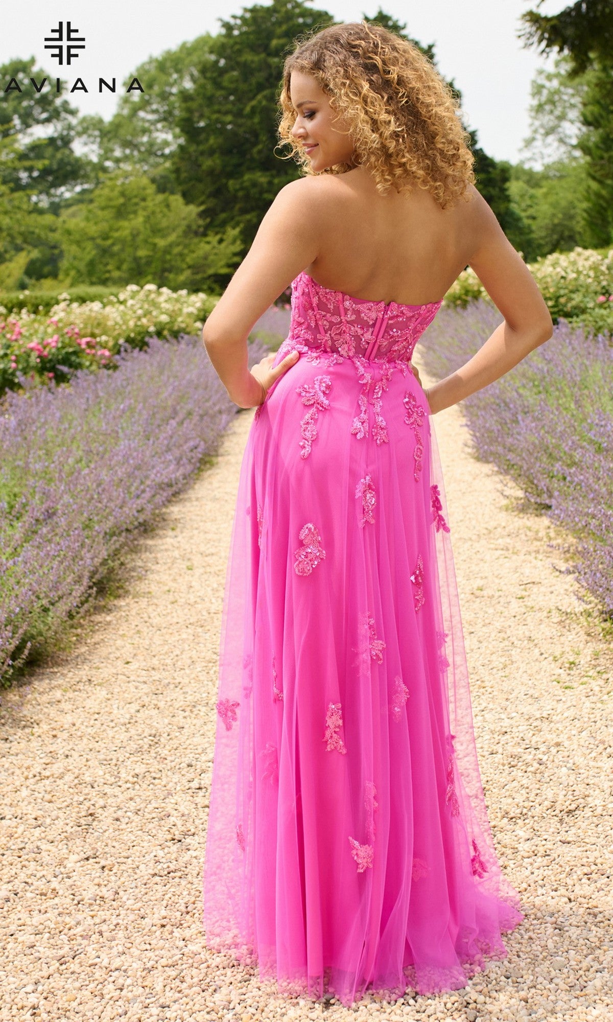 A woman stands on a gravel path in a garden, wearing the Faviana 11474 long prom dress featuring a strapless corset bodice and floral embroidery.