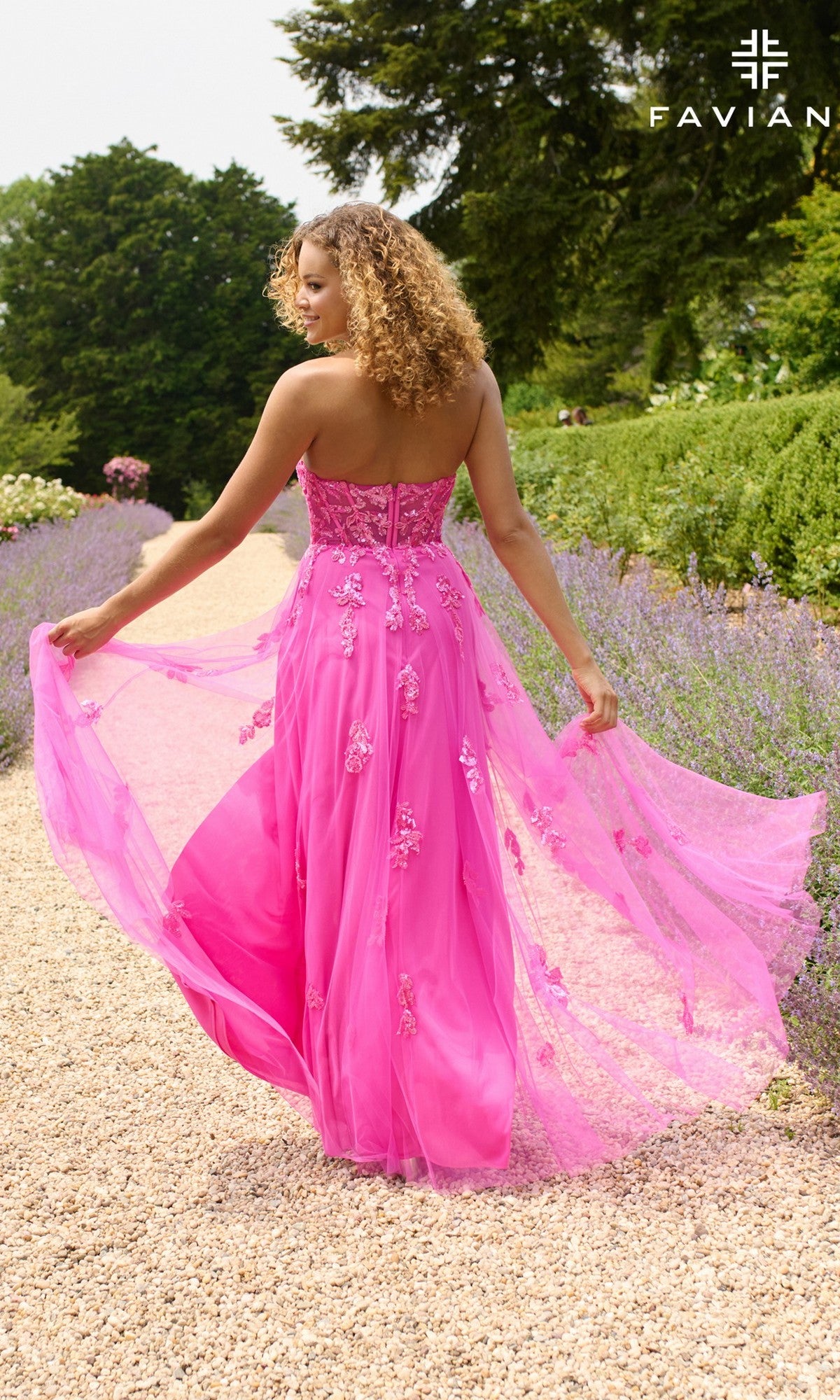 A woman with curly hair walks through a garden on a gravel path, holding out her Faviana 11474 long prom dress featuring a strapless corset bodice and bright pink embroidered details.