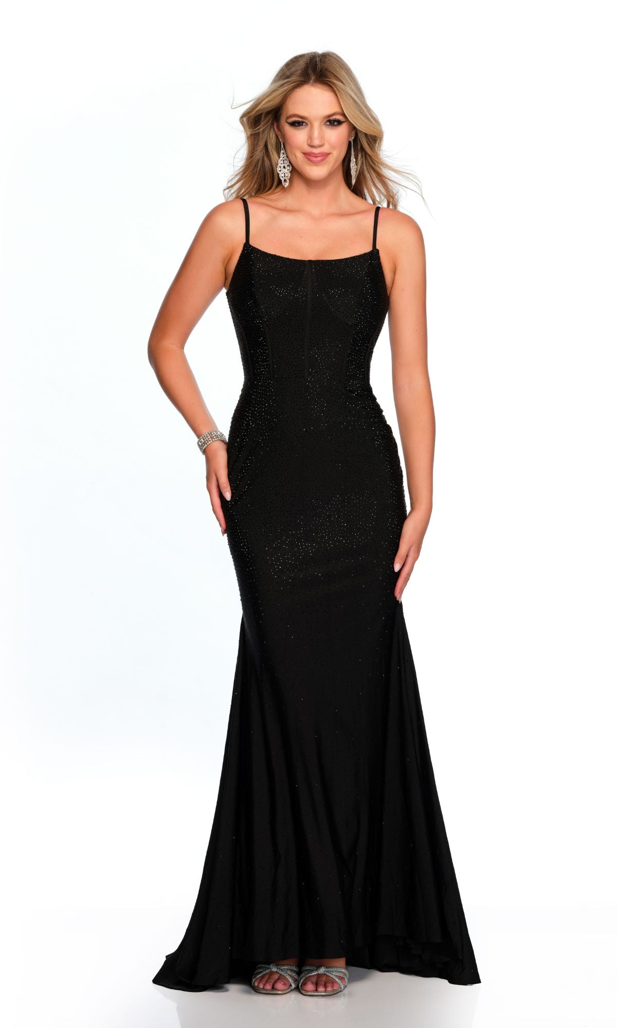 A woman wears the Long Formal Dress 11576 by Dave and Johnny—a fitted black gown with a scoop neckline and thin straps—paired with silver earrings, a bracelet, and sandals, standing against a plain white background.