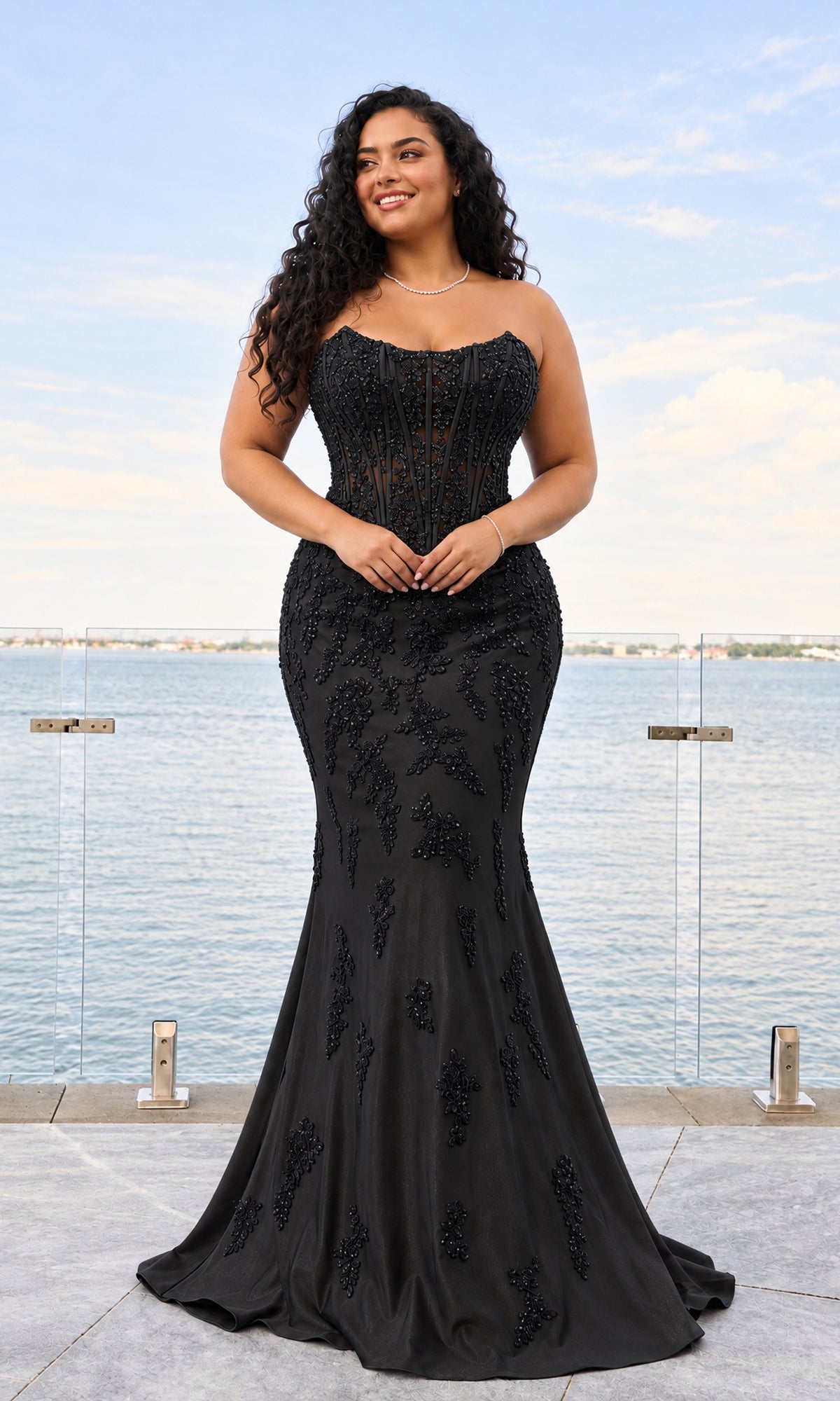 A woman in a black, strapless, bead-embroidered Long Plus-Size Prom Dress: Faviana 11600E stands on a balcony overlooking the water.