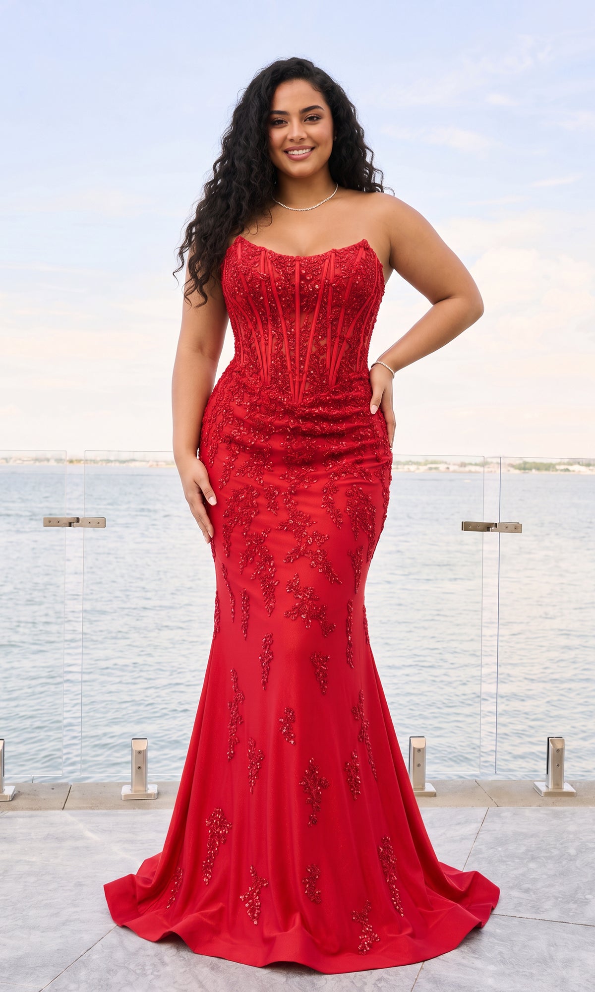 A woman wears a red, strapless Long Plus-Size Prom Dress: Faviana 11600E, a corset gown with a floor-length skirt and beaded floral embroidery.