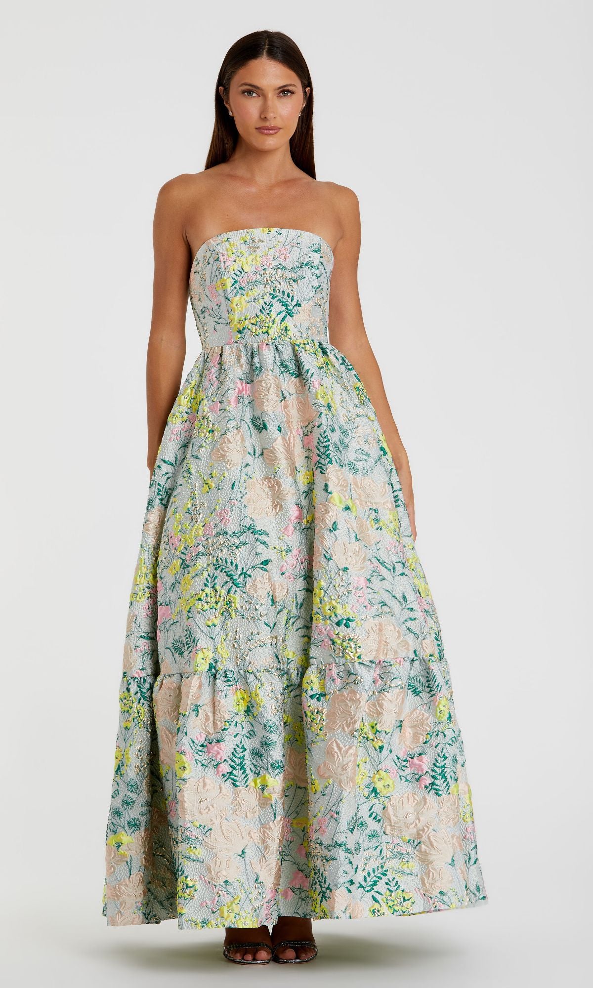 A woman stands against a plain background wearing the Strapless Brocade Formal Gown: Mac Duggal 11604, a long, strapless formal dress with a pastel multicolor floral print.