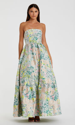 A woman stands against a plain background wearing the Strapless Brocade Formal Gown: Mac Duggal 11604, a long, strapless formal dress with a pastel multicolor floral print.