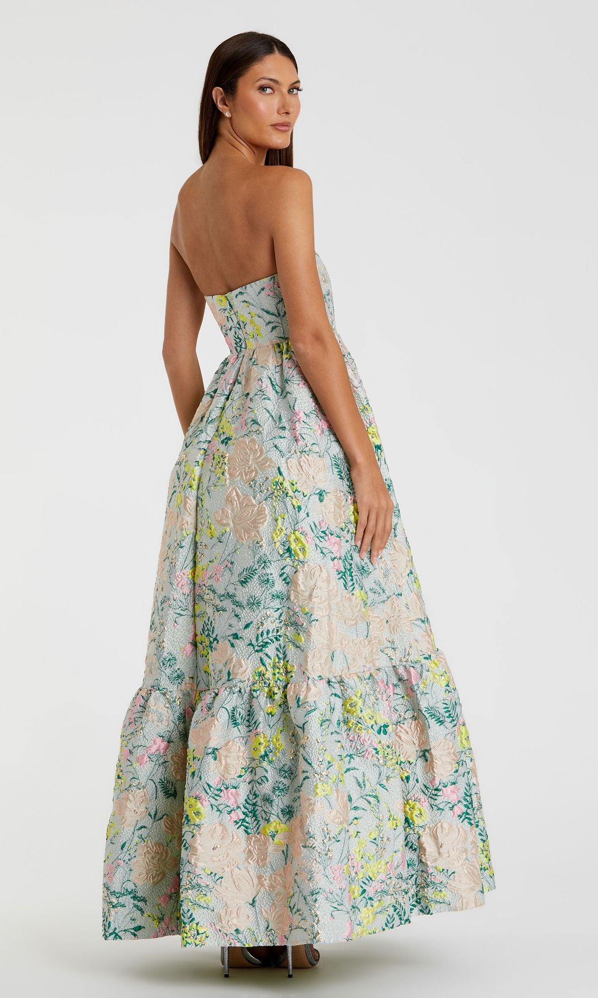 A woman wears the Mac Duggal 11604 Strapless Brocade Formal Gown—a floor-length, multicolor evening dress with a tiered skirt—standing slightly turned against a plain background.