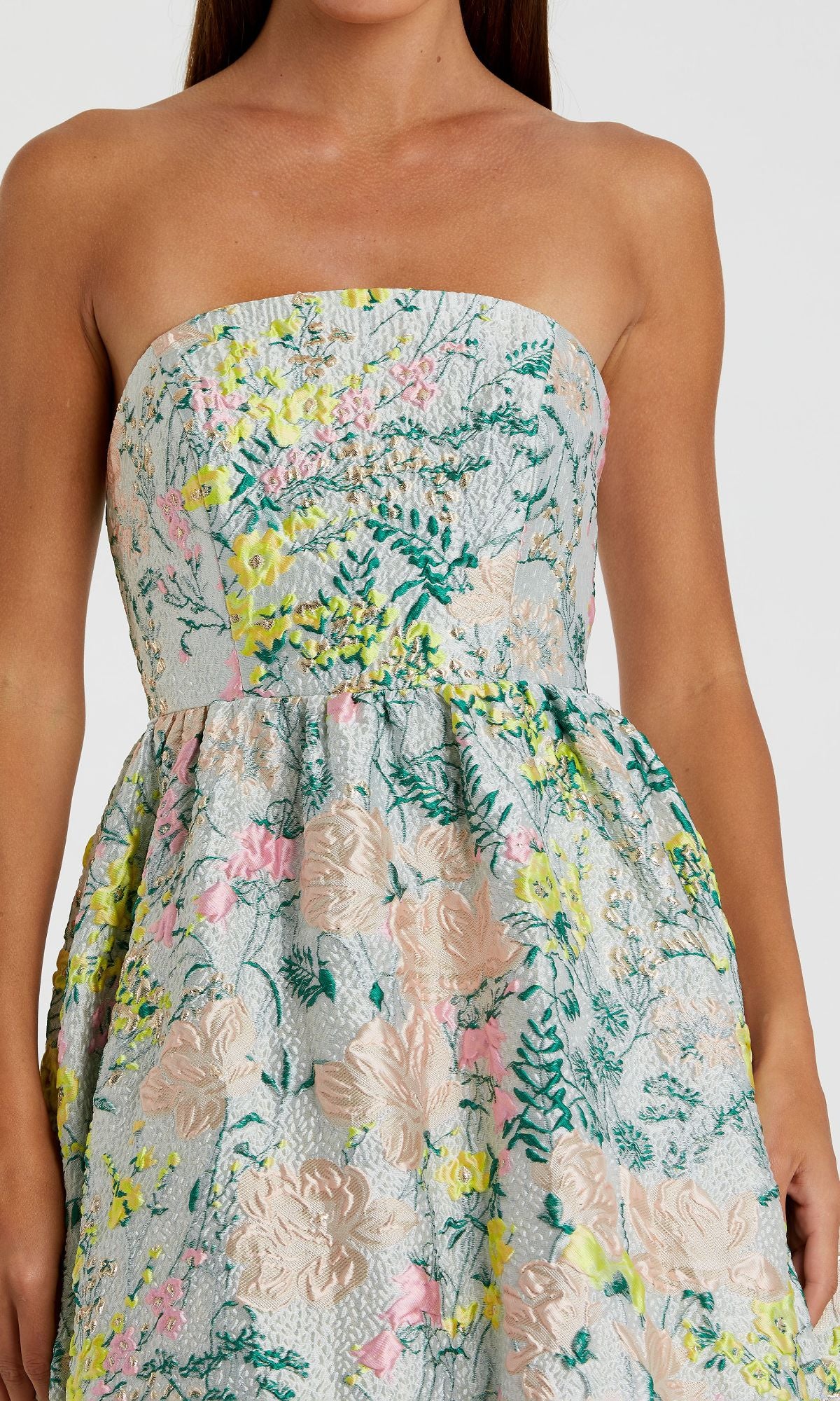 A woman wears the Strapless Brocade Formal Gown by Mac Duggal 11604, featuring a textured floral pattern in pastel yellow, pink, and green on a light background.