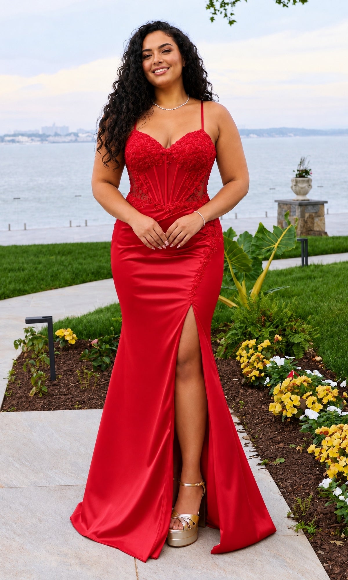 A woman wearing a red Long Plus-Size Prom Dress: Faviana 11605E with a corset bodice, thin straps, and a high side slit stands on a paved path near a body of water.