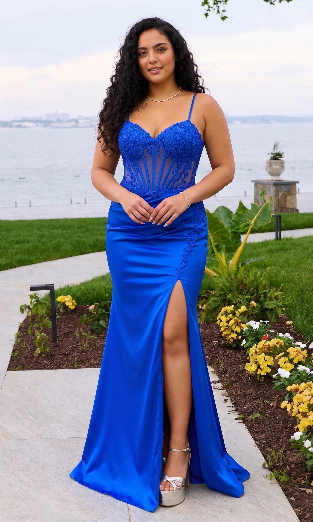 A person wears a royal blue, floor-length Faviana 11605E plus-size prom dress with a corset bodice featuring lace appliques and a high thigh slit.