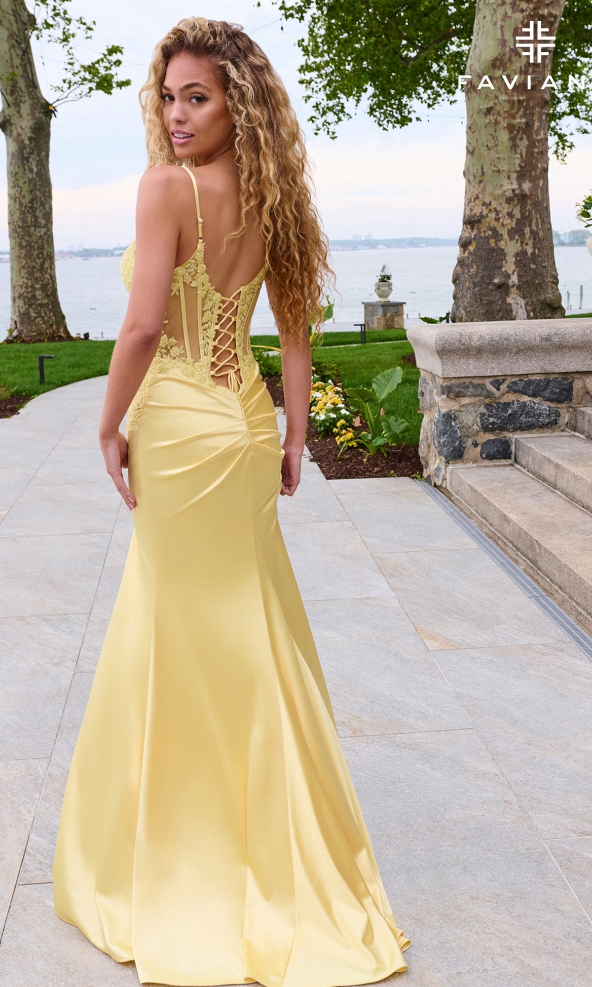 A woman wears a pale yellow, floor-length satin Long Prom Dress: Faviana 11605 featuring a sheer corset bodice and a lace-up back.