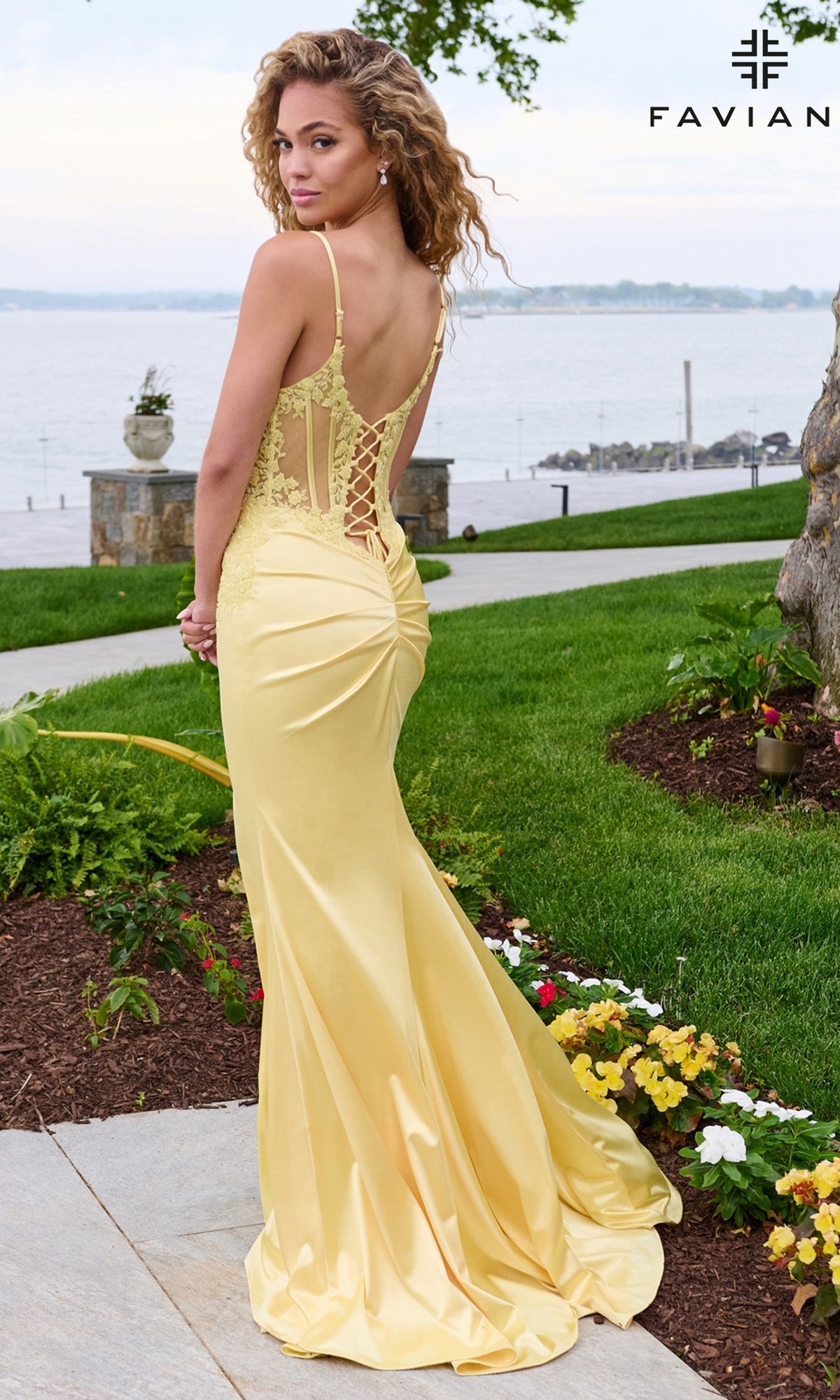 A woman stands near a waterfront wearing a yellow satin Faviana 11605 prom dress with a lace bodice and a corset-style lace-up back.