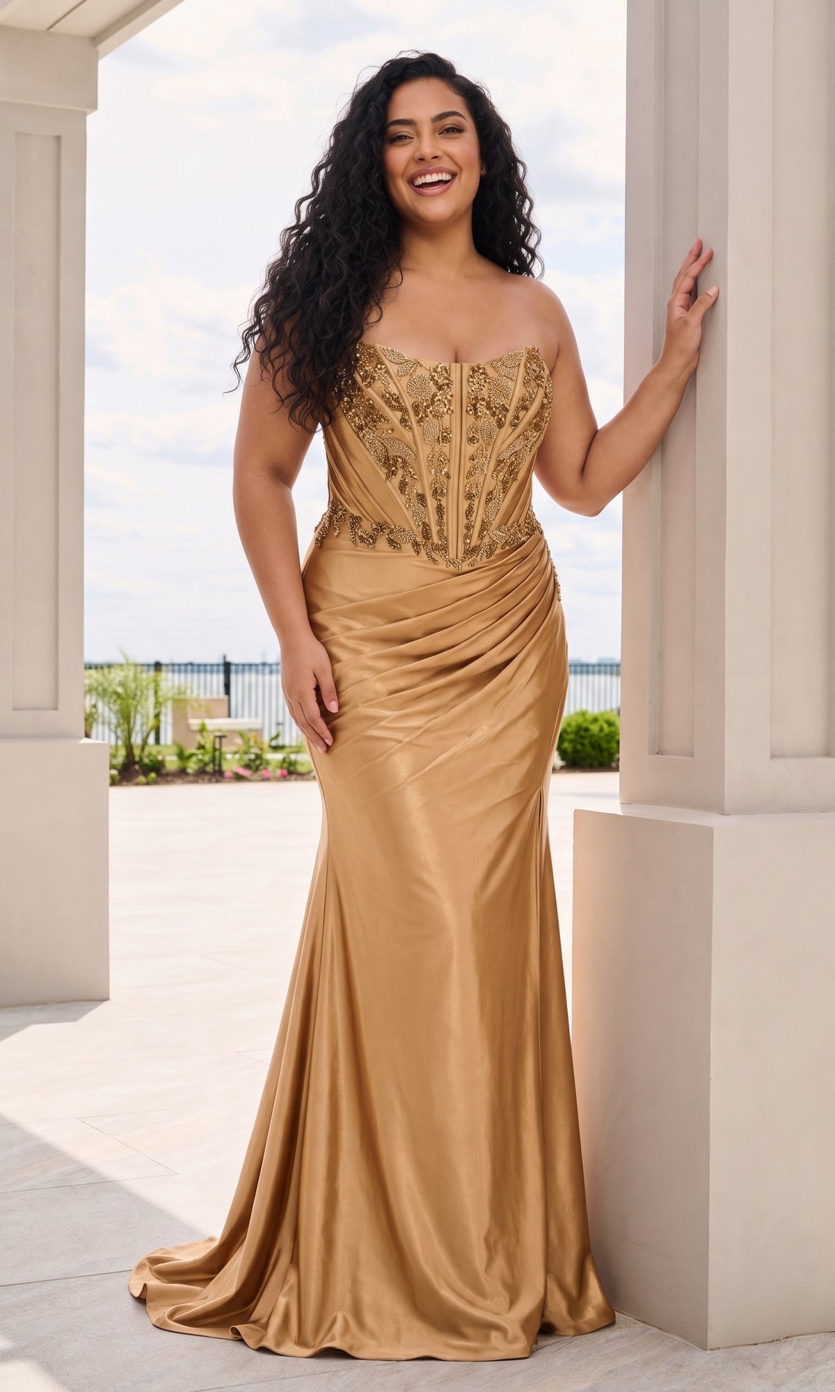 A smiling person wears a gold Long Plus-Size Prom Dress: Faviana 11606E, a strapless, floor-length satin gown featuring an intricate, beaded corset bodice and a ruched skirt.