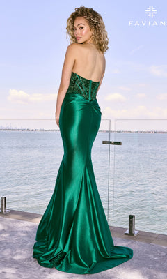 A person wears a floor-length, strapless emerald green Long Prom Dress: Faviana 11607 with a beaded corset bodice and satin skirt, standing on a balcony overlooking the water.
