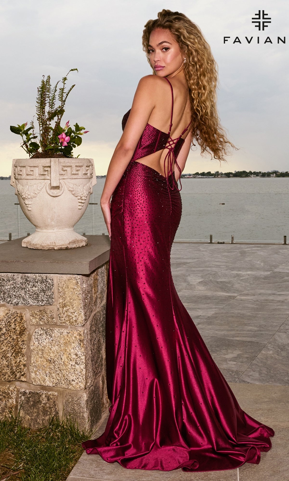 Seen from behind, a woman wears the Faviana 11621 long prom dress in maroon satin, featuring a lace-up open back and bead embellishments.