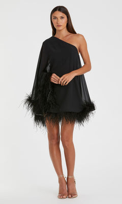 A woman models the Mac Duggal 11623 short wedding guest dress, featuring a black one-shoulder silhouette with a feather-trimmed hem, styled with nude heels—an elegant choice against a white backdrop.