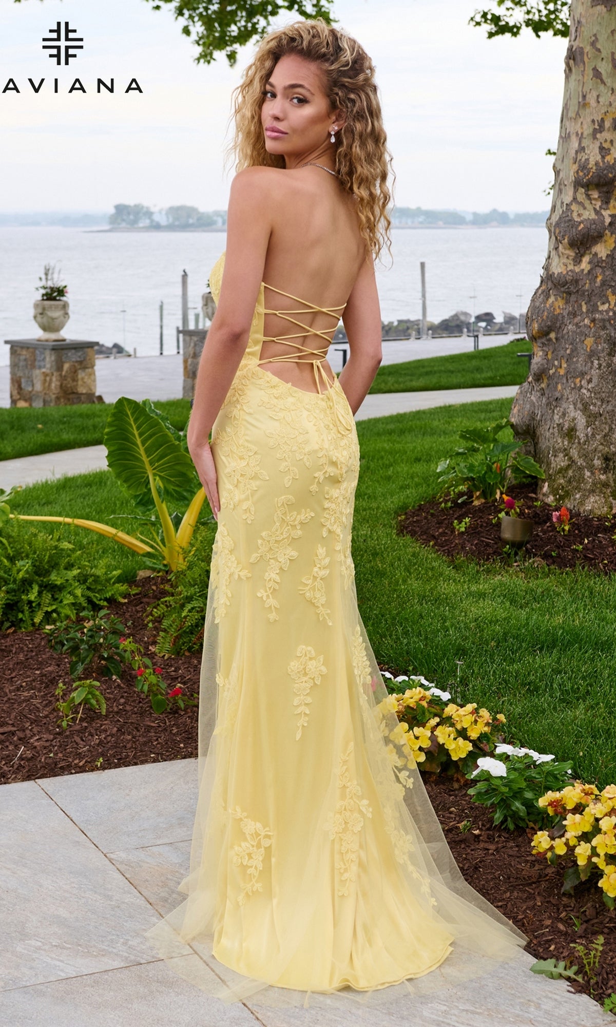 A woman wearing the pale yellow, floor-length strapless Faviana 11627 column prom dress with an open, lace-up back and intricate lace appliques.