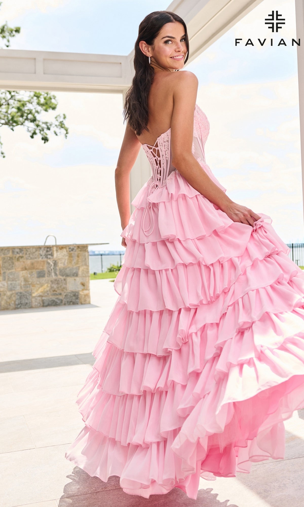A woman wears a light pink, floor-length Faviana 11629 tiered prom dress featuring a strapless corset bodice and a ruffled skirt.