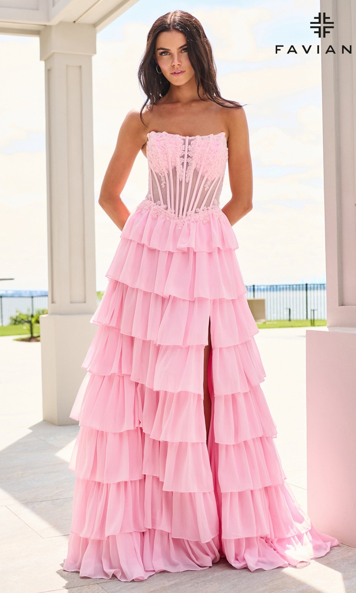 A woman wears the light pink Faviana 11629 long prom dress, a strapless, floor-length gown with a corset bodice, a tiered chiffon ruffle skirt, and a high leg slit.