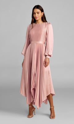A woman models the Mac Duggal 11638 Fuchsia Midi Wedding Guest Dress, featuring long sleeves, pleats, and an asymmetrical hem—ideal for wedding guests—styled with beige heels against a plain gray background.