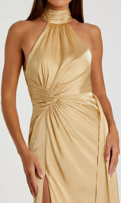 A woman in a Mac Duggal 11642 high-neck, sleeveless gold satin prom dress features ruched waist detailing and a side slit.