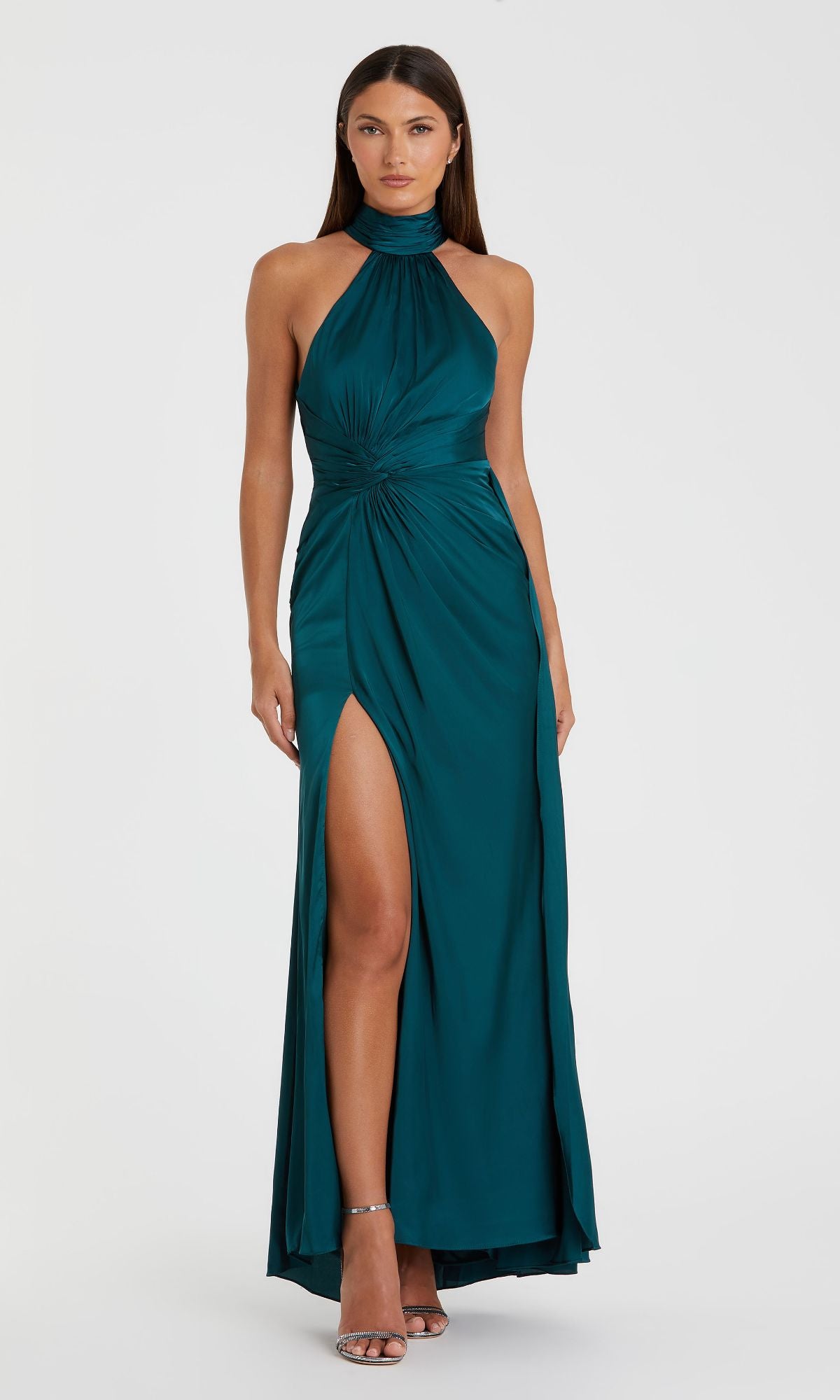 A woman stands against a plain light background wearing the Mac Duggal 11642 High-Neck Long Prom Dress in teal, featuring a halter neck, high slit, and gathered knot waist detail.