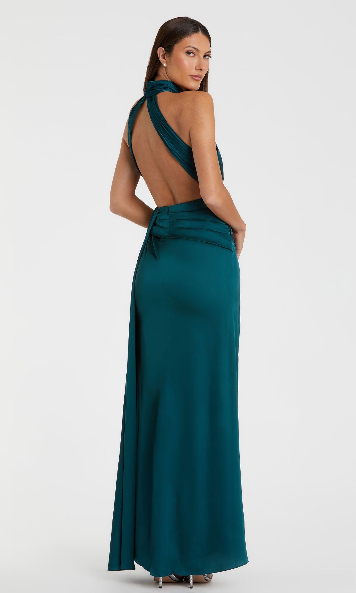 A woman poses with her back to the camera in the High-Neck Long Prom Dress Mac Duggal 11642, a teal floor-length gown with a halter neckline, set against a plain white background.