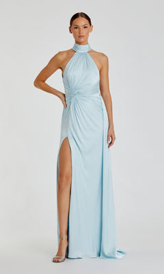A woman poses elegantly in the Mac Duggal 11642 High-Neck Long Prom Dress—a light blue gown with halter neck, high slit, gathered waist—and silver high-heeled sandals against a plain background.
