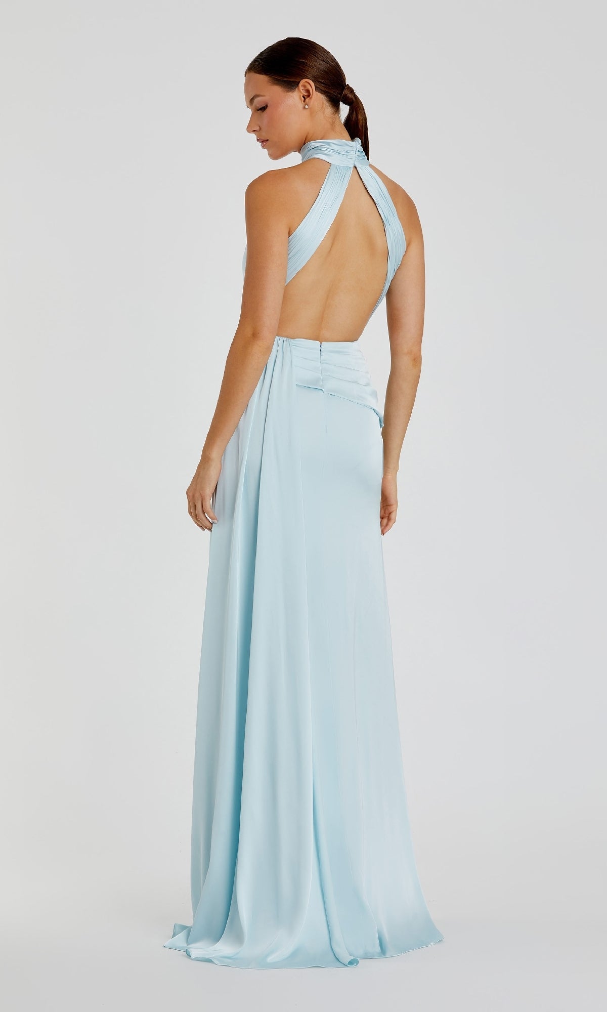 A woman models the Mac Duggal 11642 High-Neck Long Prom Dress in light blue, featuring a backless halter neckline, as she stands facing slightly away against a plain background.