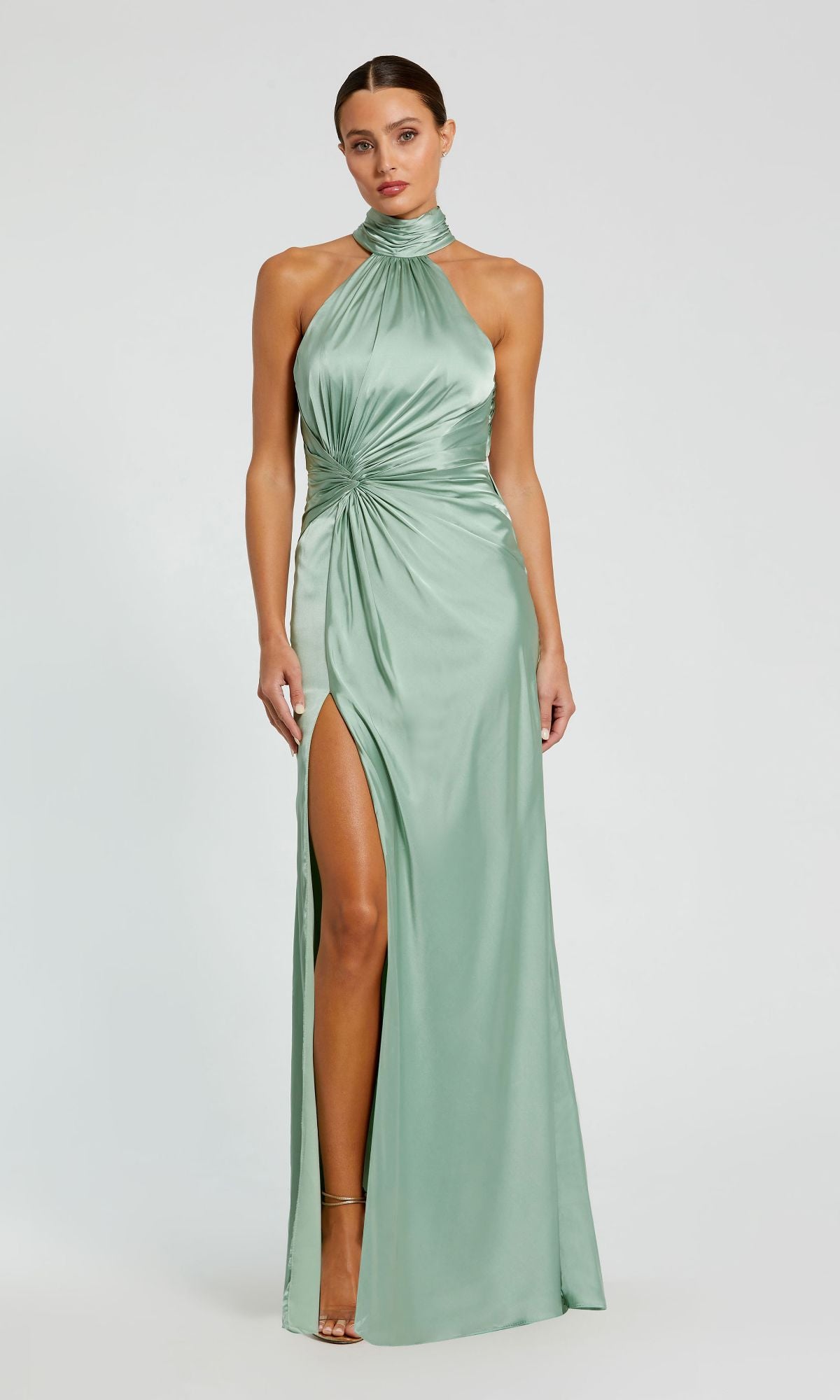 A woman stands against a plain light background wearing the Mac Duggal 11642 High-Neck Long Prom Dress in light green satin, featuring a ruched waist detail and a high slit.