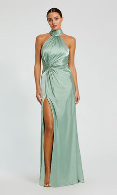 A woman stands against a plain light background wearing the Mac Duggal 11642 High-Neck Long Prom Dress in light green satin, featuring a ruched waist detail and a high slit.