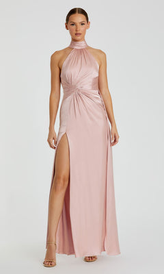 A woman wears the Mac Duggal 11642 High-Neck Long Prom Dress in blush pink, featuring a sleeveless design, high slit, ruched waist, and pairs it with nude heels.