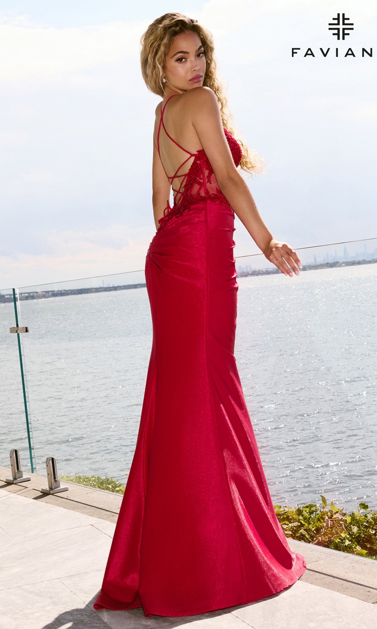 Woman wearing a red Long Prom Dress: Faviana 11646 mermaid gown with a beaded lace bodice and strappy open back, standing on a terrace overlooking the water.